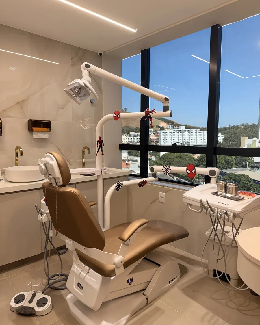 Consultório com cadeira odontológica decorada com enfeites de super-herói e janela panorâmica para os prédios e morros da cidade.