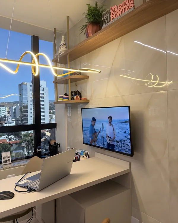 Consultório com mesa de atendimento, televisão na parede e janela para os prédios da cidade.